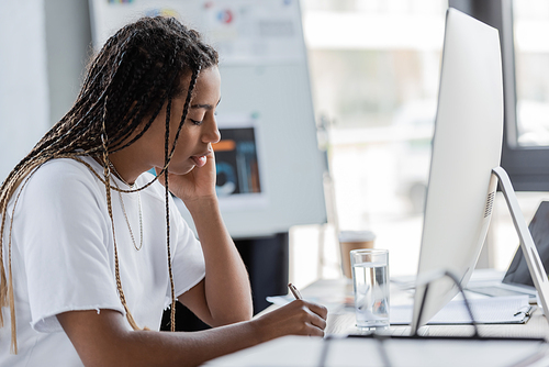 African american businesswoman in t-shirt writing near computer and glass of water in office