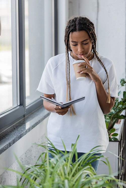 African american businesswoman holding coffee to go and notebook near plants in office