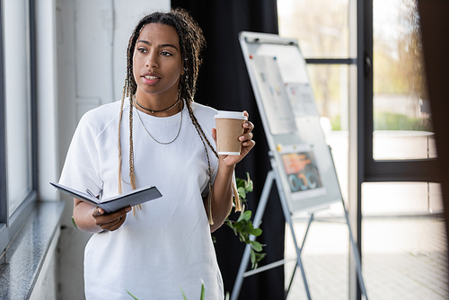 African american businesswoman holding notebook and takeaway drink in office