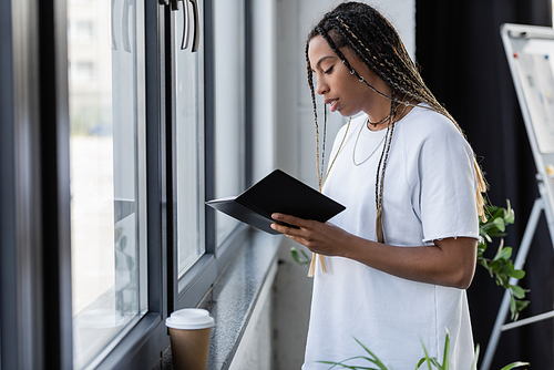 Young african american businesswoman holding notebook near coffee to go on windowsill in office