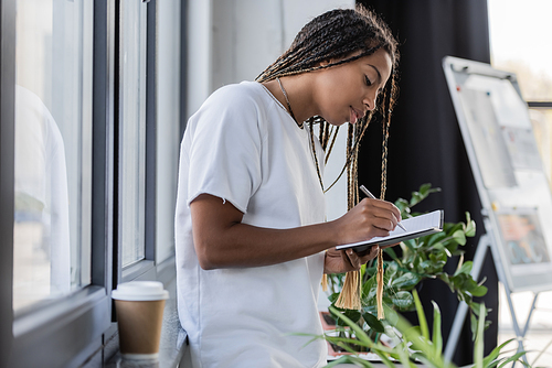 African american businesswoman writing on notebook near blurred coffee to go on windowsill in office