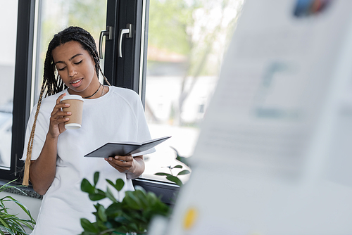 Smiling african american businesswoman holding paper cup and notebook in office