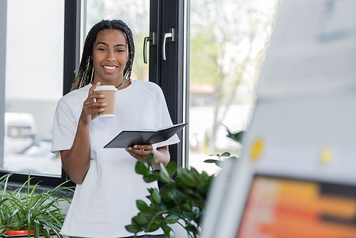 Positive african american businesswoman in t-shirt holding takeaway drink and notebook in office