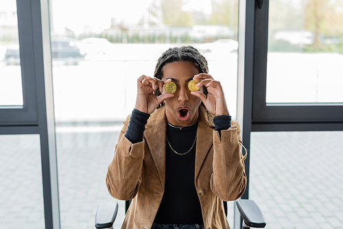 KYIV, UKRAINE - APRIL 27, 2022: Shocked african american businesswoman holding bitcoins near eyes in office