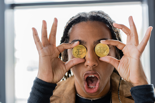 KYIV, UKRAINE - APRIL 27, 2022: Shocked african american businesswoman holding bitcoins near eyes in office