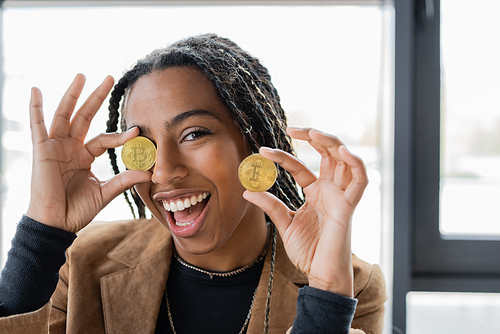 KYIV, UKRAINE - APRIL 27, 2022: Cheerful african american businesswoman holding bitcoins in office