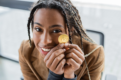 KYIV, UKRAINE - APRIL 27, 2022: Smiling african american businesswoman holding bitcoin and looking at camera