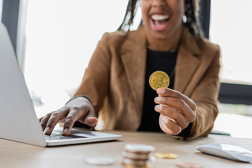 KYIV, UKRAINE - APRIL 27, 2022: Cropped view of blurred african american businesswoman holding bitcoin near laptop in office