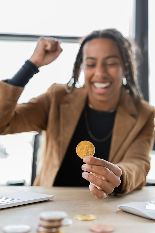 KYIV, UKRAINE - APRIL 27, 2022: Blurred excited african american businesswoman holding bitcoin near laptop in office