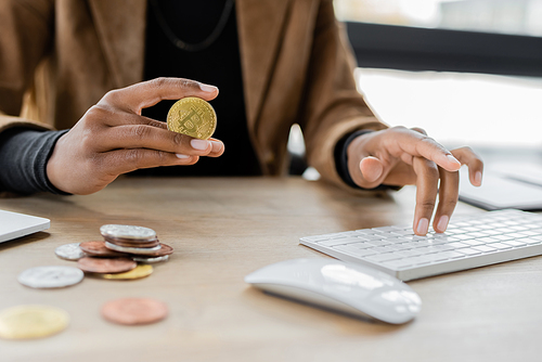 KYIV, UKRAINE - APRIL 27, 2022: Cropped view of african american businesswoman holding bitcoin and typing on computer keyboard