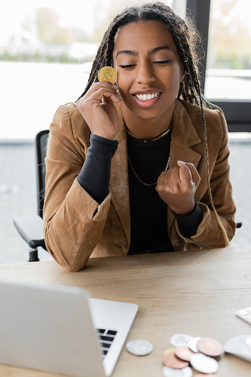 KYIV, UKRAINE - APRIL 27, 2022: Excited african american businesswoman holding bitcoin near laptop in office