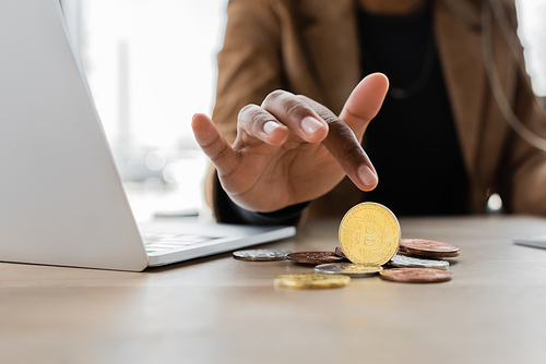 KYIV, UKRAINE - APRIL 27, 2022: cropped view of african american businesswoman pointing at bitcoins near laptop in office