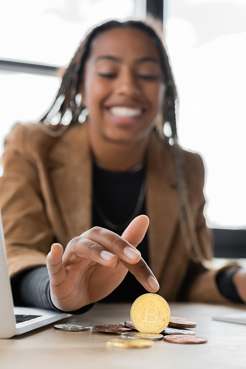 KYIV, UKRAINE - APRIL 27, 2022: Blurred african american businesswoman touching bitcoin near laptop in office