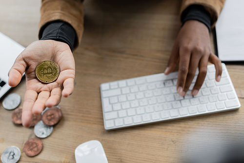 KYIV, UKRAINE - APRIL 27, 2022: Cropped view of african american businesswoman holding bitcoin and typing on computer keyboard