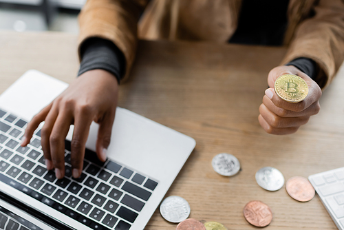 KYIV, UKRAINE - APRIL 27, 2022: Cropped view of african american businesswoman using laptop and holding bitcoin in office