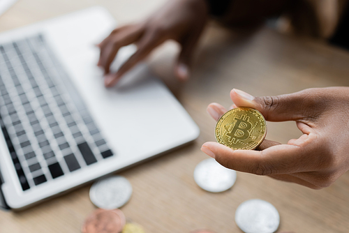 KYIV, UKRAINE - APRIL 27, 2022: Cropped view of african american businesswoman holding golden bitcoin and using laptop in office