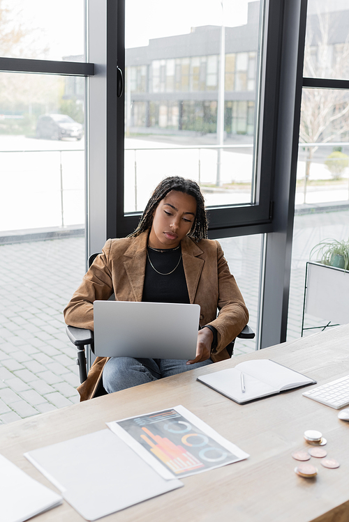 KYIV, UKRAINE - APRIL 27, 2022: African american businesswoman in jacket using laptop near papers and notebook in office