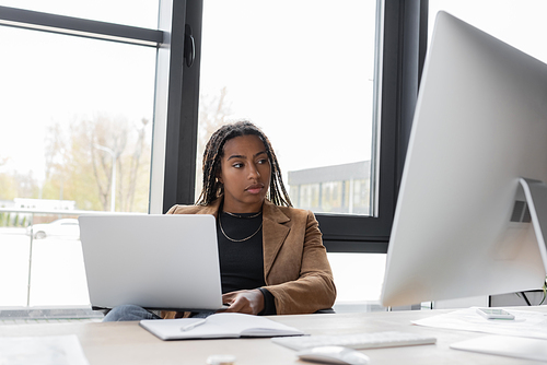 African american businesswoman in jacket holding laptop near computer monitor in office