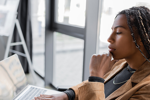 Side view of african american businesswoman using blurred laptop with blank screen in office