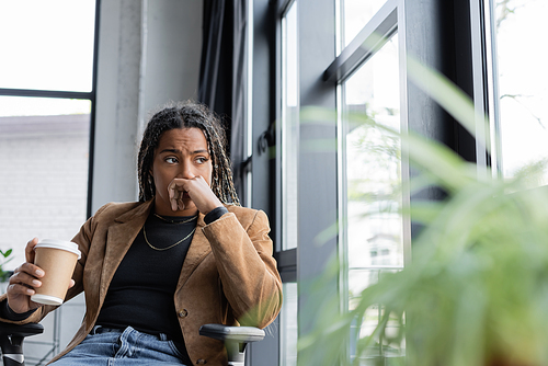 Worried african american businesswoman in blazer holding takeaway drink near window in office