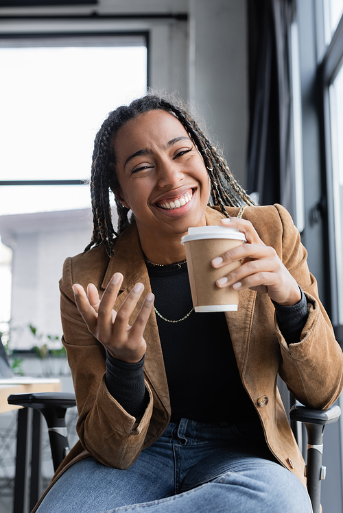 Positive african american businesswoman in jacket holding paper cup and looking at camera in office