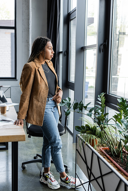 Side view of african american businesswoman in jacket and jeans looking at window in office