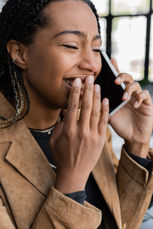 Cheerful african american businesswoman in blazer talking on smartphone in office