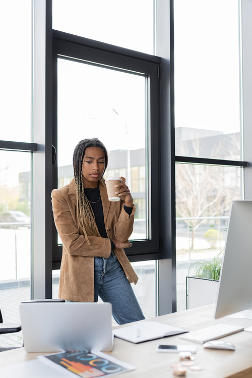 African american businesswoman holding coffee to go near devices and notebook in office