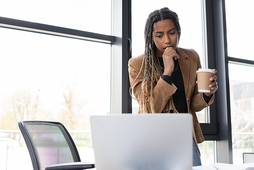 Focused african american businesswoman holding coffee to go and looking at laptop in office