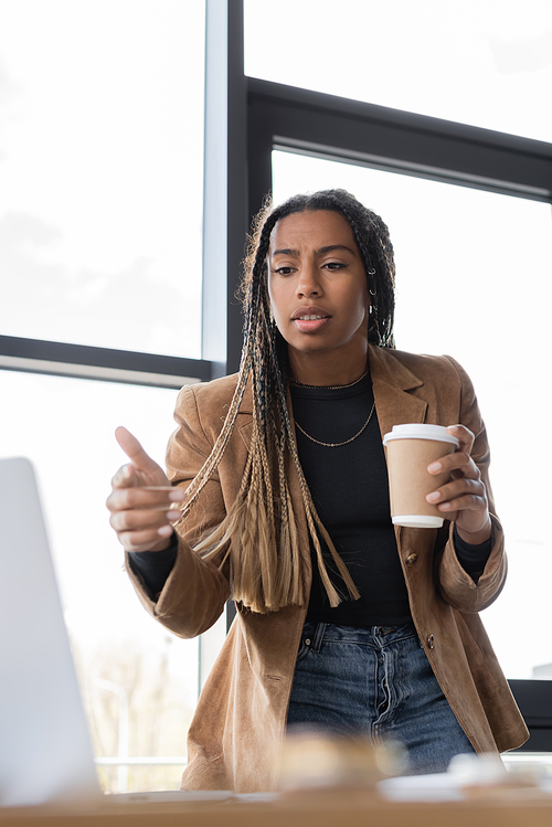 African american businesswoman holding coffee to go and pointing with finger at laptop in office