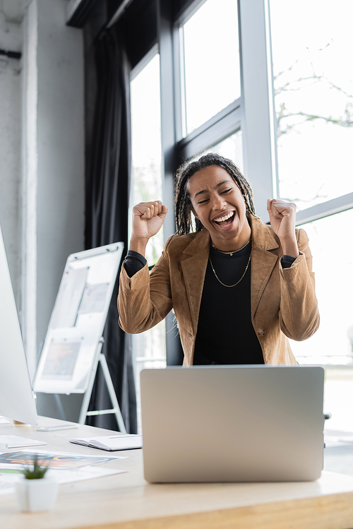 Excited african american businesswoman looking at laptop near papers on table in office