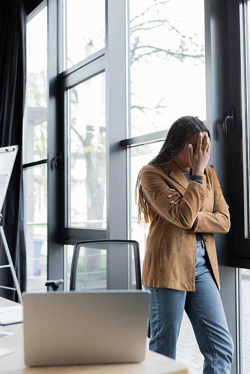 Upset african american businesswoman standing near window and blurred laptop in office