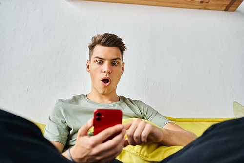 surprised student in his 20s at home sitting on yellow couch and scrolling in social media