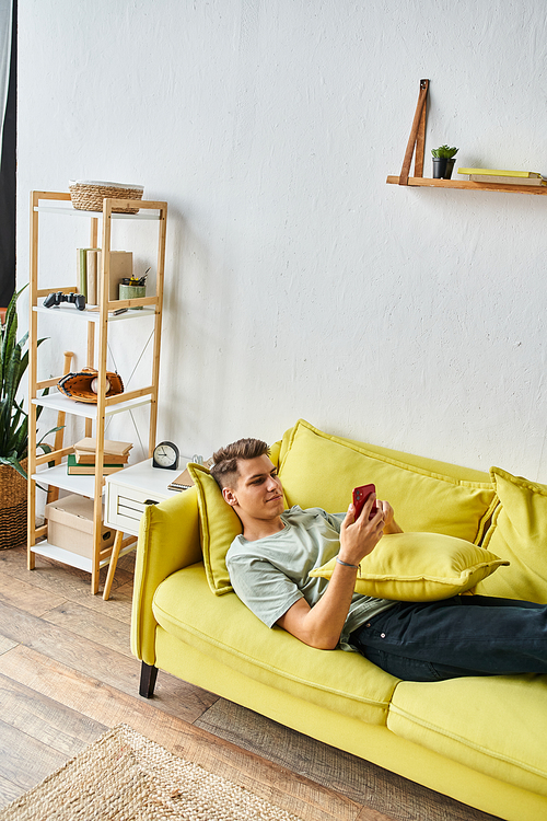 charming young man lying on yellow couch in living room and scrolling to social media