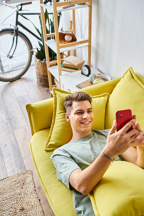 smiling young student lying on yellow couch in living room and scrolling to social media
