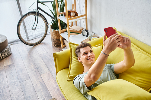 attractive young man with brown hair lying on yellow couch in living room and taking selfie