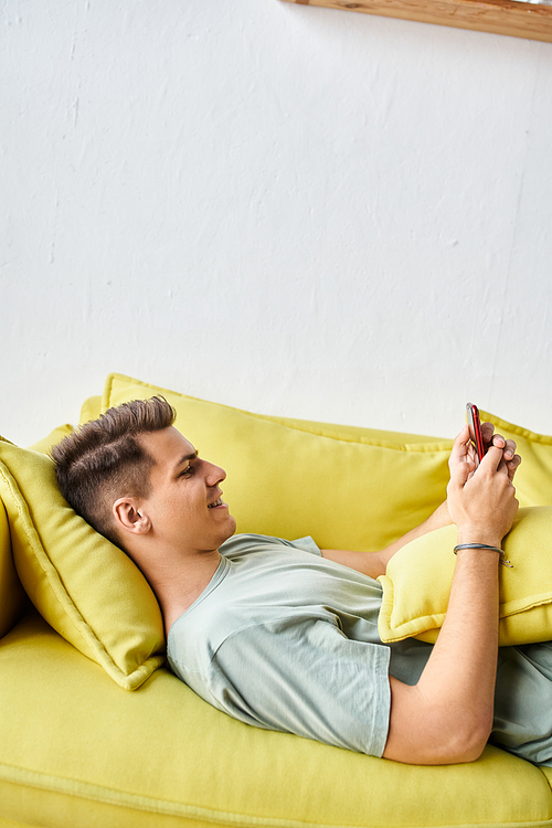 profile of cheerful student lying on yellow couch in living room and scrolling to social media