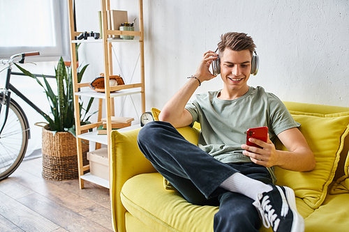 smiling young guy in headphones sitting on yellow couch at home and scrolling to social media