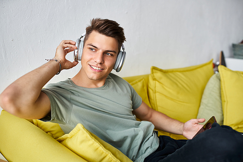 charismatic man with headphones and smartphone in yellow couch at home looking to window