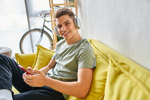 smiling man in headphones in yellow couch at home texting on smartphone and looking to camera