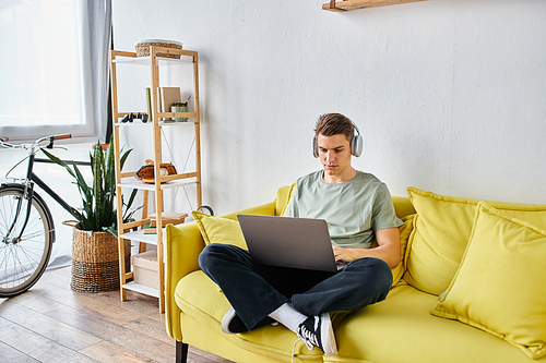 focused student with brown hair in headphones in yellow couch at home learning on laptop