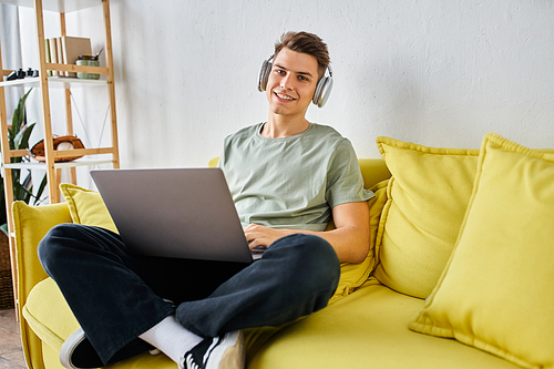 handsome young man with headphones and laptop in yellow couch at home looking to camera
