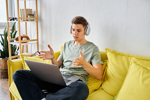 confident student with headphones in yellow couch at home speaking to online meeting in laptop