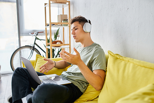 focused student with headphones in yellow couch at home speaking to online meeting in laptop