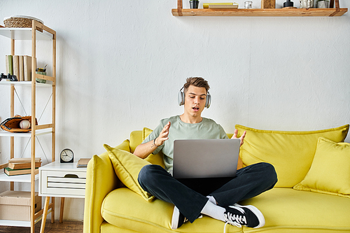 surprised student with headphones and laptop in yellow couch at home speaking to online meeting