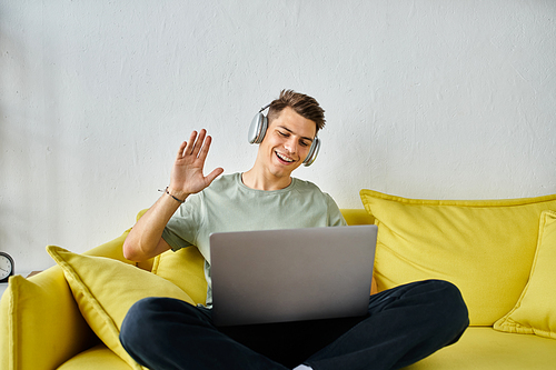 handsome young man with headphones and laptop in yellow couch saying hello to online meeting
