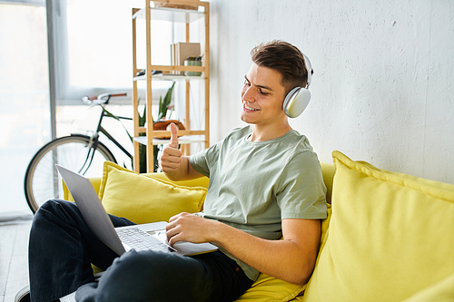 smiling young man with headphones and laptop in yellow couch liking to online meeting