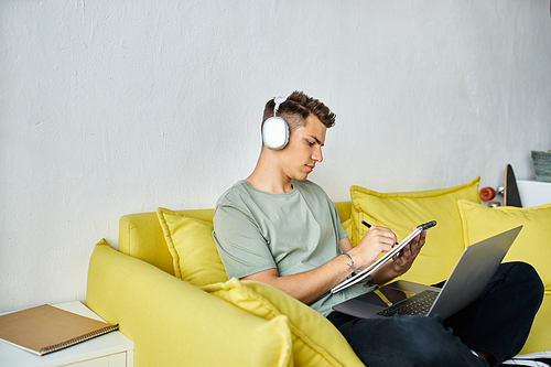 focused student with headphones and laptop in yellow couch studying and writing in note