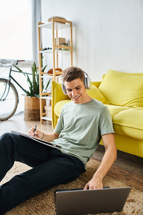 cheerful guy with headphones on floor near yellow couch studying in laptop and writing in note