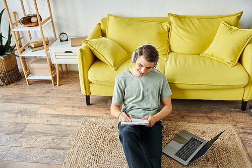 smiling guy with headphones and laptop on floor near yellow couch studying and writing in note